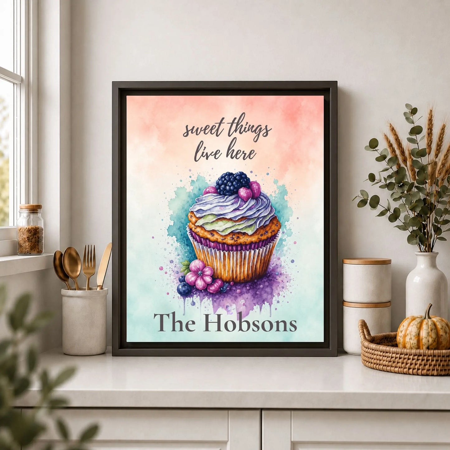 Framed artwork of a colorful cupcake with 'sweet things live here' text on a kitchen counter.