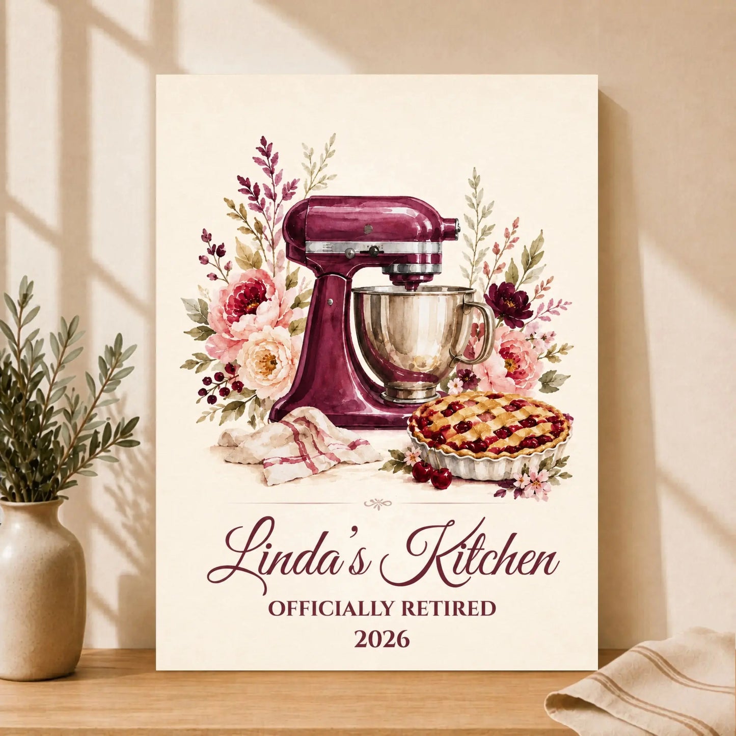 Framed artwork of a kitchen scene with a mixer, pie, and flowers, featuring ' Officially Retired 2026'.